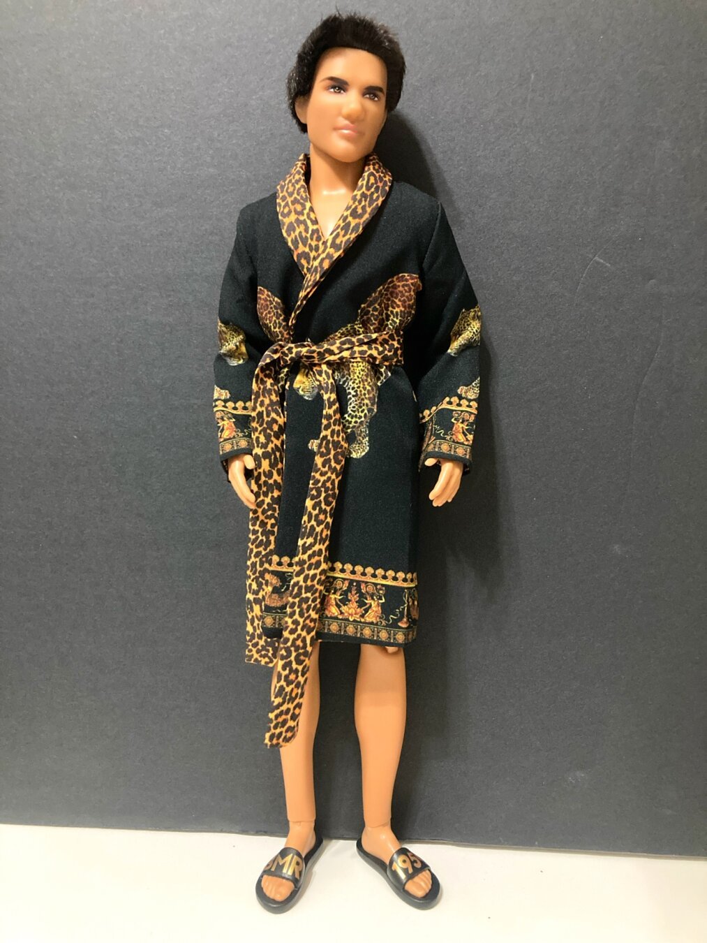 Jacob from Twilight Made to Move hybrid Ken doll is posed standing on a white surface against a black backdrop. He wears a black bathrobe featuring images of cheetahs on each side and cheetah-print collar and belt, along with black slide sandals printed with “BMR” and “1959”, respectively, on the viewer’s left and right.