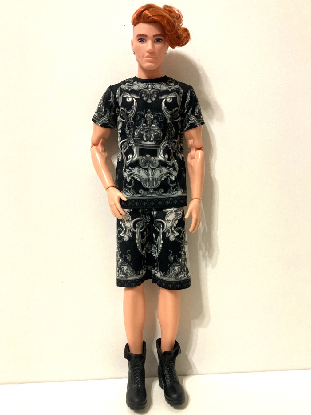BMR1959 Ken doll with red hair standing centered against a white background wearing a black t-shirt and shorts combo with elaborate grey baroque-inspired scrollwork printed on both pieces to mimic designer wear. He completes the look with open combat boots.