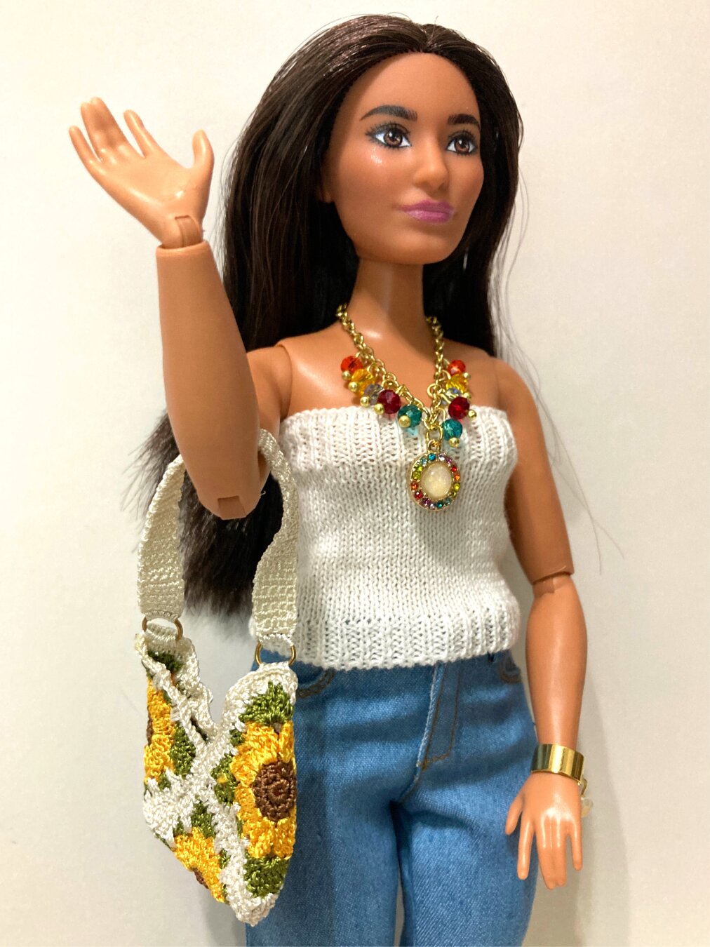 A brunette medium-complected curvy articulated hybrid with the head of Barbie fashionista 200 stands against a white background. She is waving, gaze fixed off to the right of the frame. She wears a white micro-knit strapless top with a colorful golden necklace and light jeans. She carries a white micro-crochet bag with a sunflower motif on the waving arm and wears a golden bangle bracelet with a pendant on the other wrist.