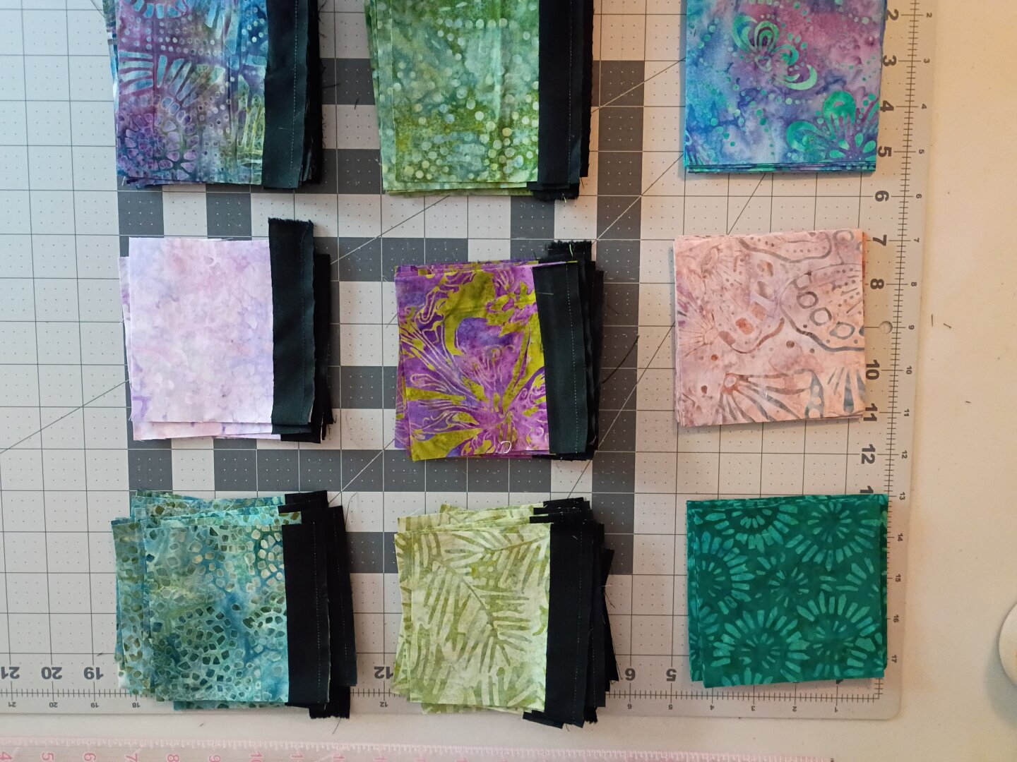 9 4-inch squares of batik fabric in shades of green and purple and pink. 6 of the squares have black sashing sewn on one side. They are laid out in a 3x3 grid to be sewn together into a 9 patch.