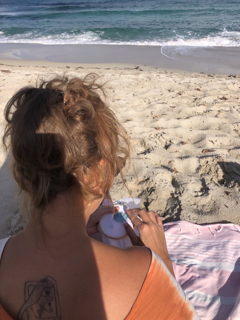 Photo où on me voit de dos (épaules et tête), assise sur une plage de sable en train de broder un petit coquillage.