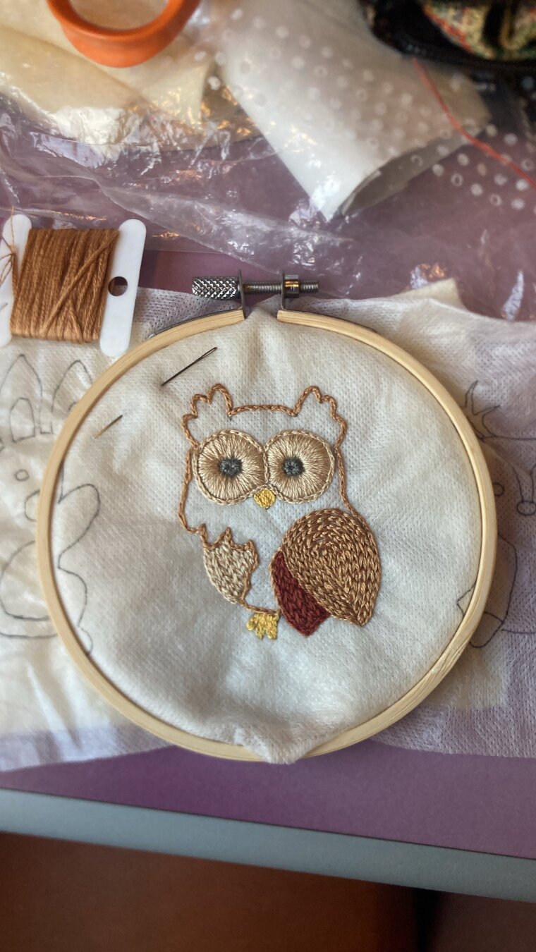 Photo d’une petite chouette mignonne en partie brodée sur de la feutrine beige. Elle est dans un tambour à broder posé sur une tablette de TGV au milieu d’un bazar d’ustensiles de broderie. On peut apercevoir un bout de ciseaux, une bobine de fil marron, du papier, un reste de feutrine.