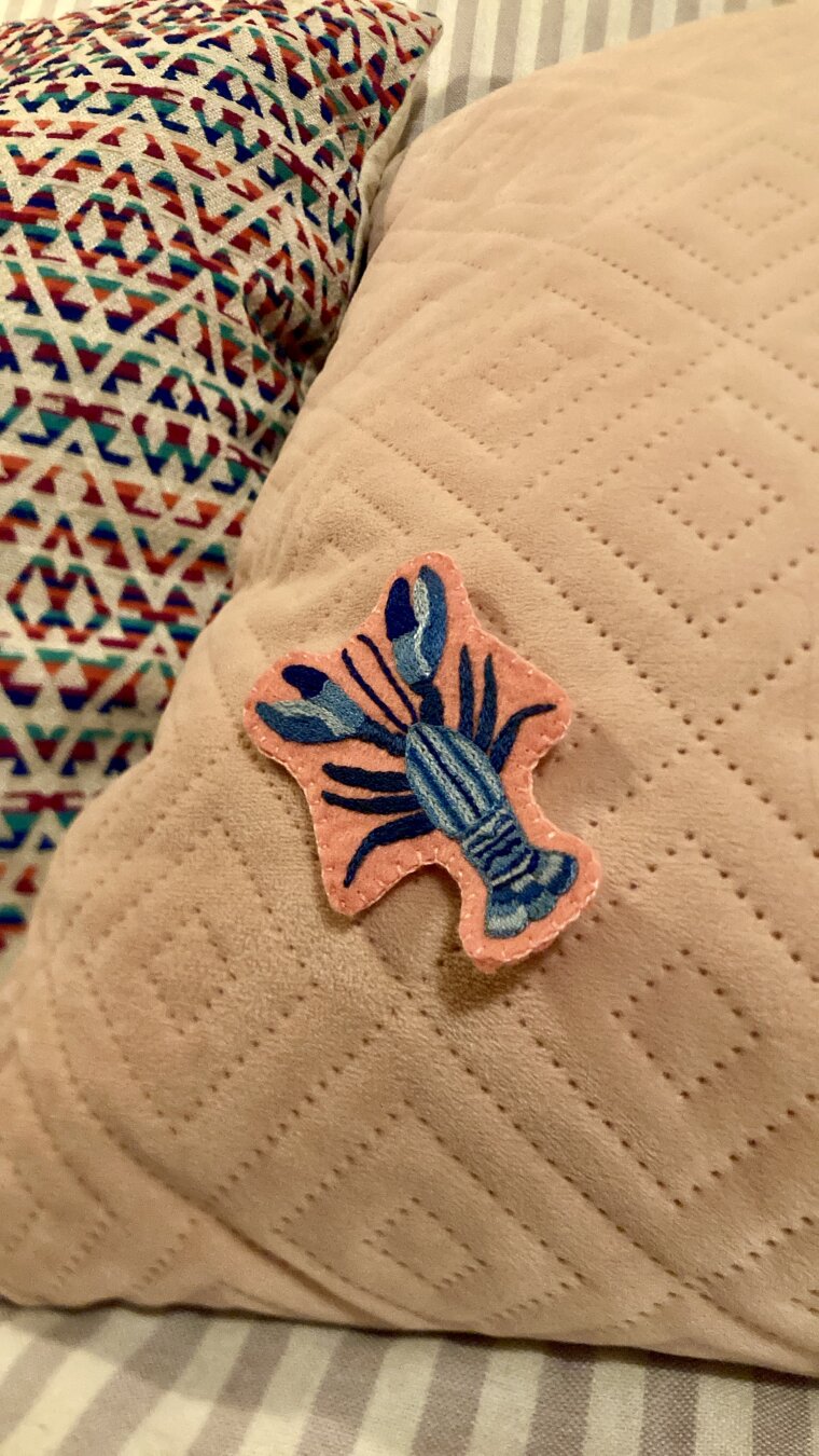 Un homard bleu brodé sur de la feutrine rose est posé sur un coussin en velouté rose pâle, on aperçoit un bout de coussin bariolé à gauche et un plaid à rayures blanches et violettes