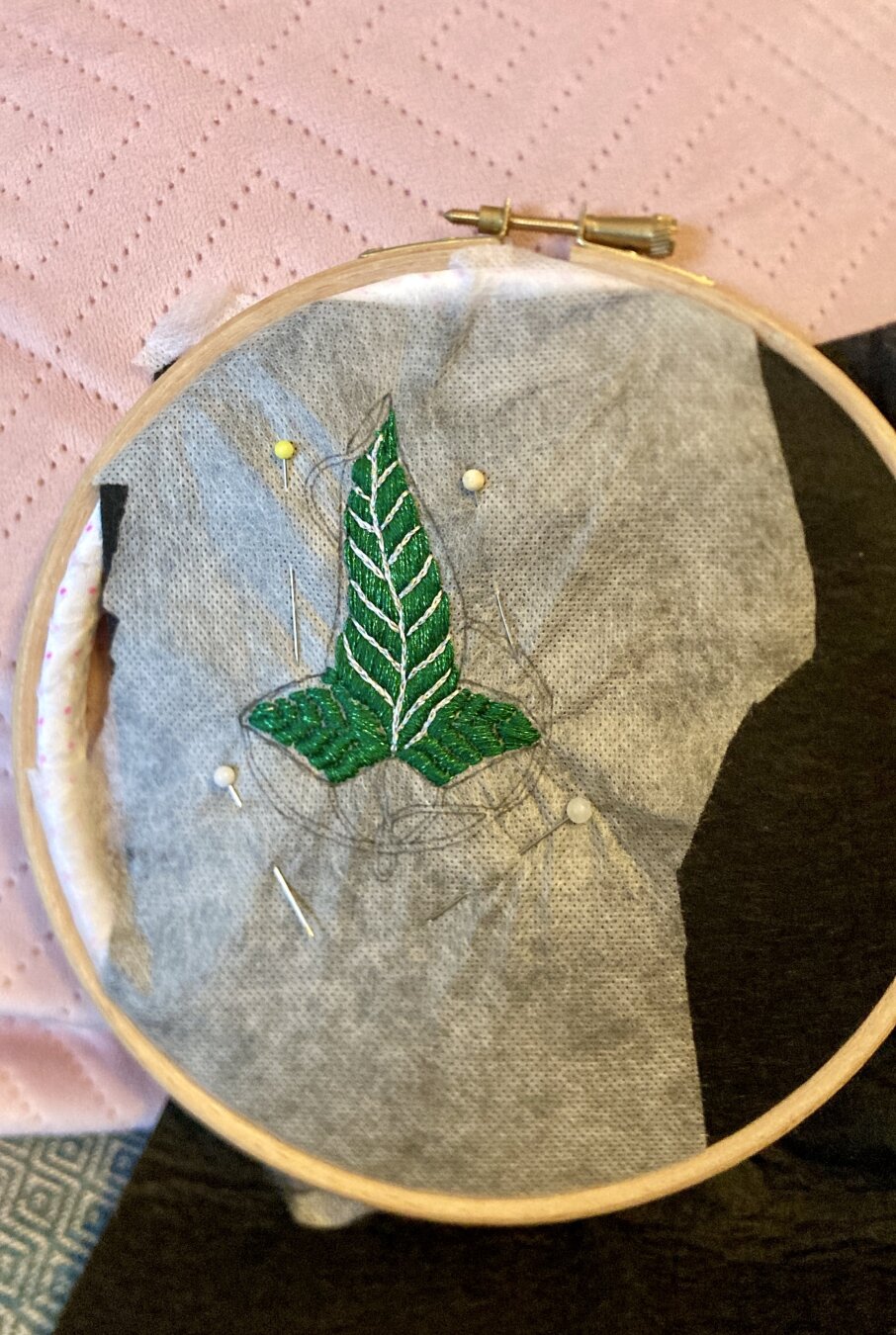 Un cercle à broder repose contre un coussin rose pâle. À l’intérieur, de la feutrine noire recouverte de tissu soluble sur lequel est brodée en partie une feuille de la Lorien en fils vert légèrement brillant et argenté.