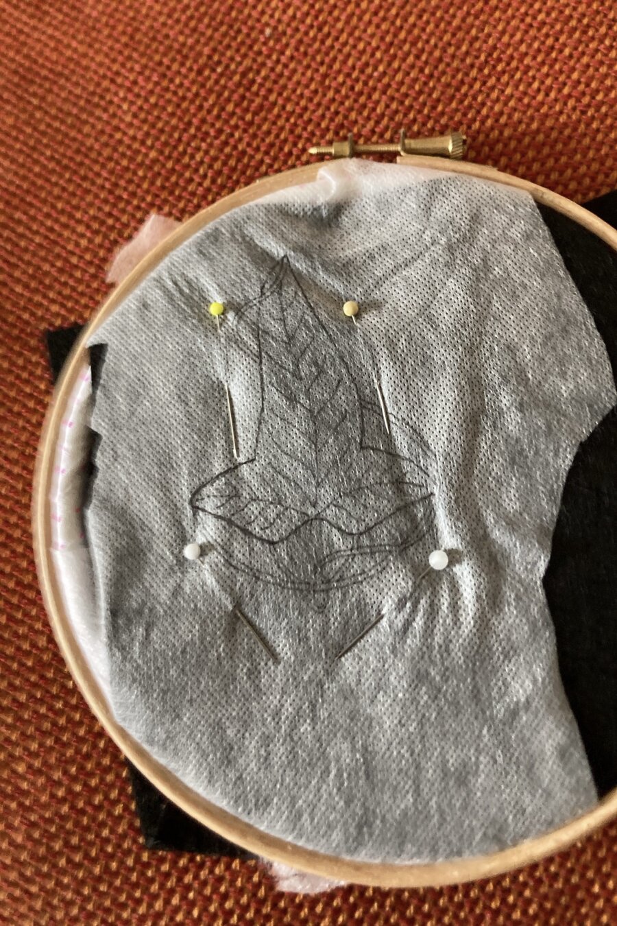 Un cercle à broder est poser sur un tissu orangé. Dans le cercle est fixée de la feutrine noire recouverte de tissu soluble. Sur le tissu est dessinée la feuille de la Lorien.