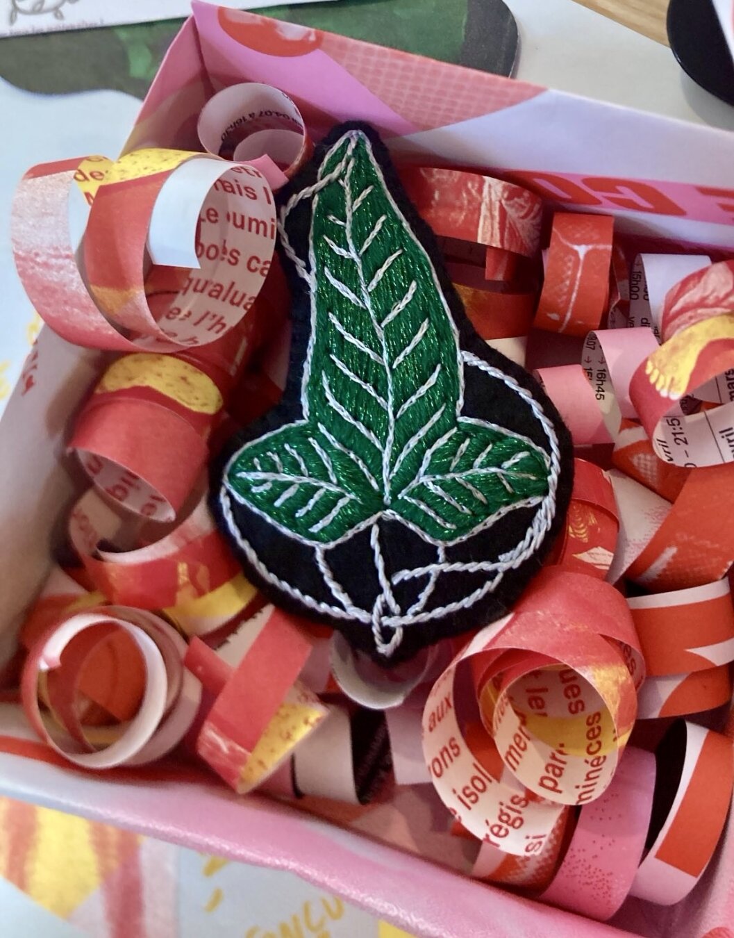 Dans une boîte en papier remplie de cotillons est posée une broche brodée représentant la feuille de la Lorien. Le fond de la broche est noir, la feuille est brodée de fils vert brillant et argenté.