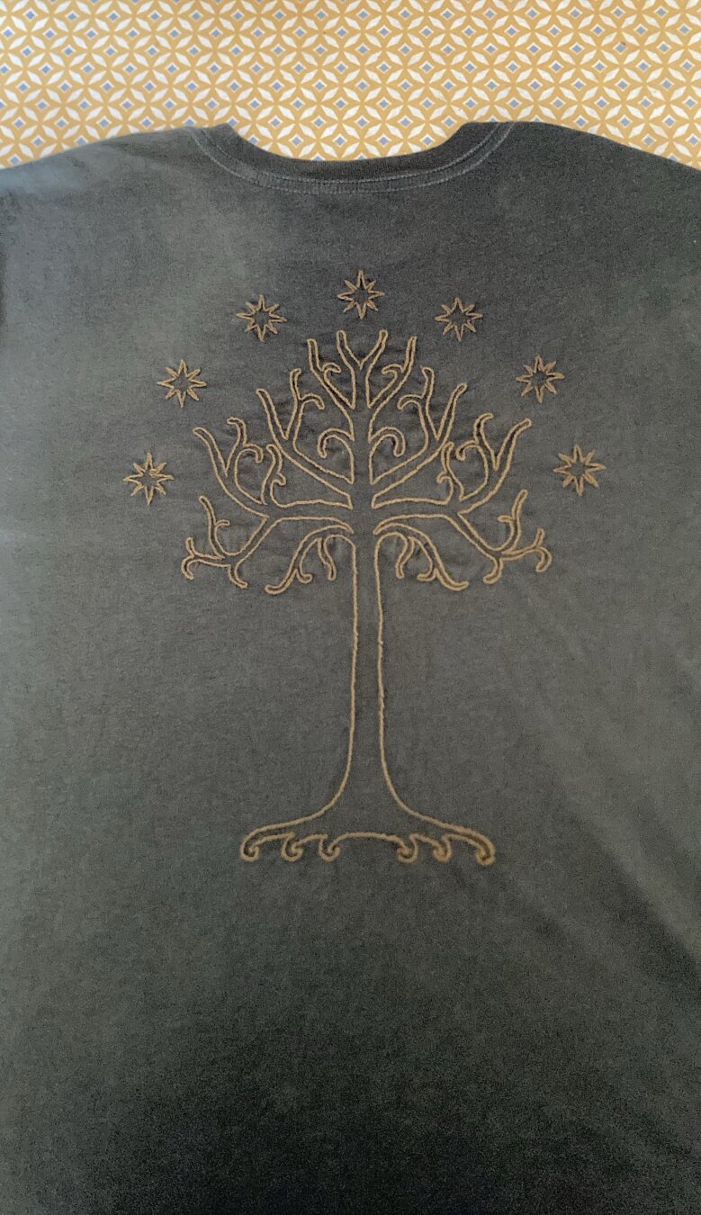 Un t-shirt vert kaki posé dos apparent sur lequel est brodé l’arbre du Gondor en ocre.