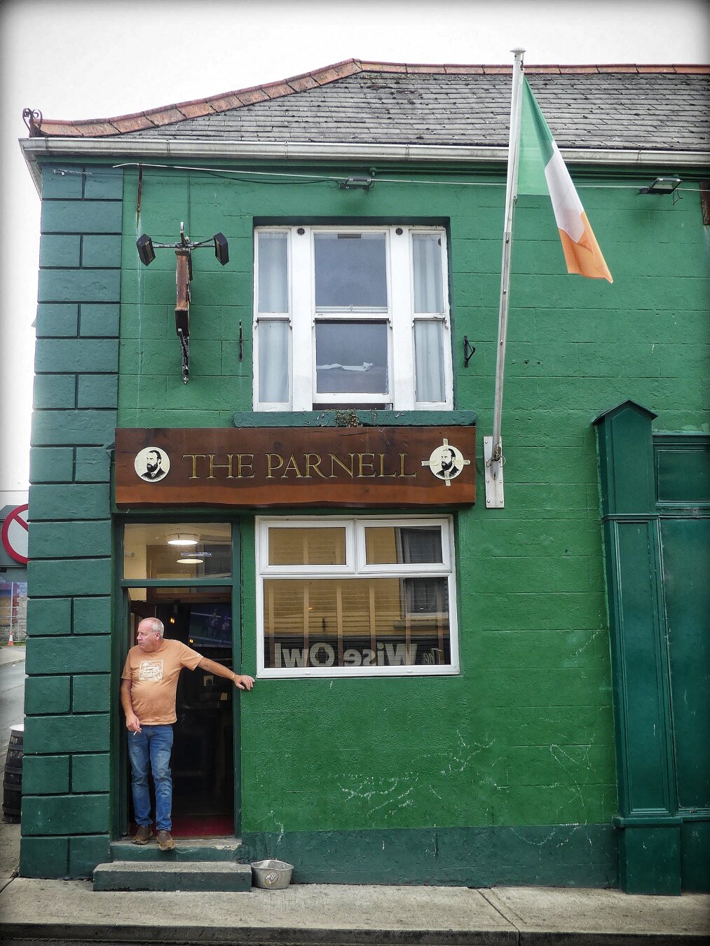 Un homme fumant une cigarette se tient devant la porte ouverte d’un pub en Irlande, il regarde la circulation des voitures