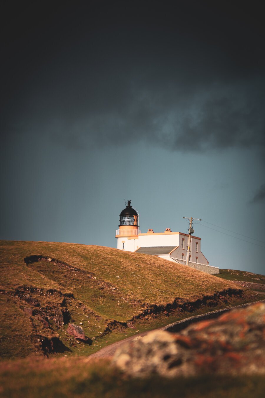 Dark blue sky at the top of the photo. The sky gets lighter below. A white lighthouse is partially hidden by brown fields and tiny hills.