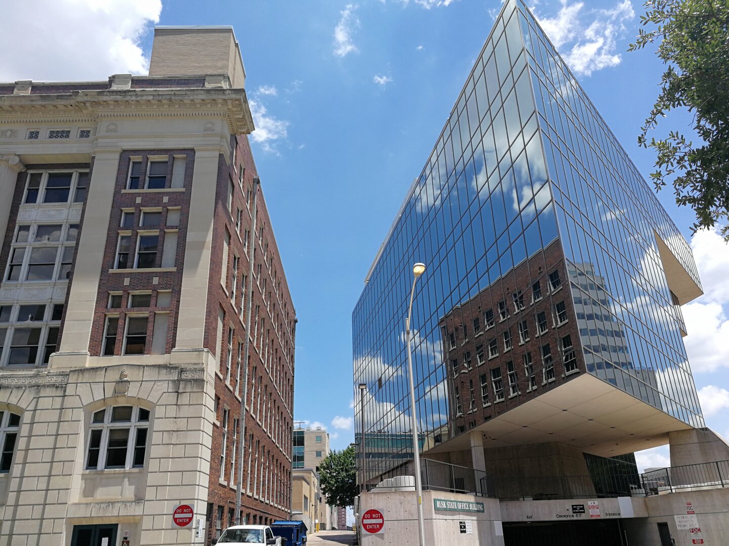 Old brick building beside modern glass building reflecting brick and sky; street with “Do Not Enter” signs and a few trees; partly cloudy day.