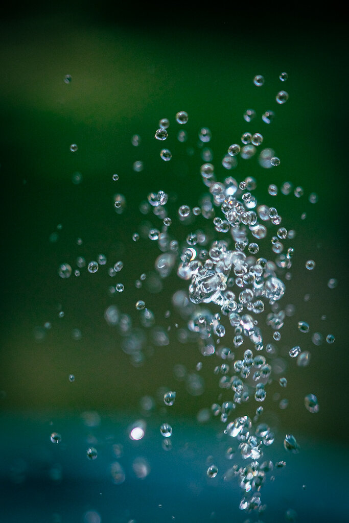 Water droplets shooting up out of a water fountain. The background is green with blue at the bottom of the frame.