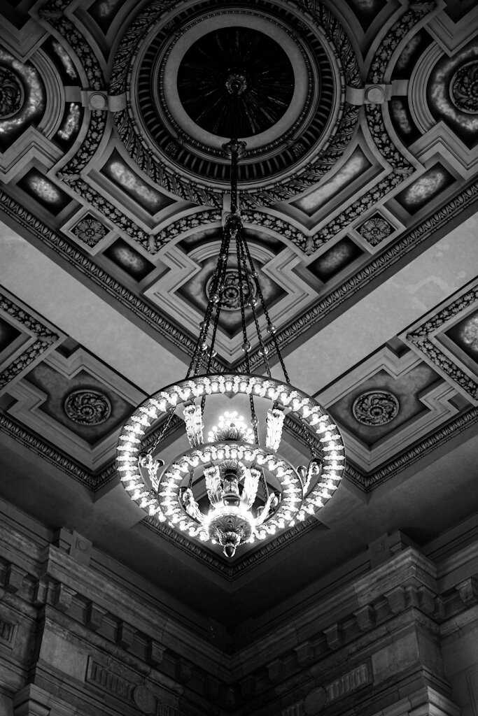 Symmetrical Black and White shot of a corner of Union Station’s ceiling in Kansas City. A large chandelier hangs down by chains and has its lights on. The ceiling has geometrical shapes and patterns across it.