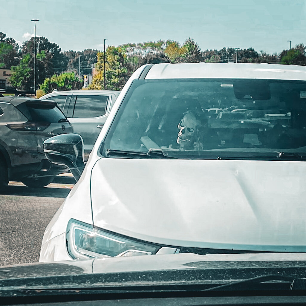 Square photo of a white vehicle in a parking lot with a fake skeleton in the passenger seat.