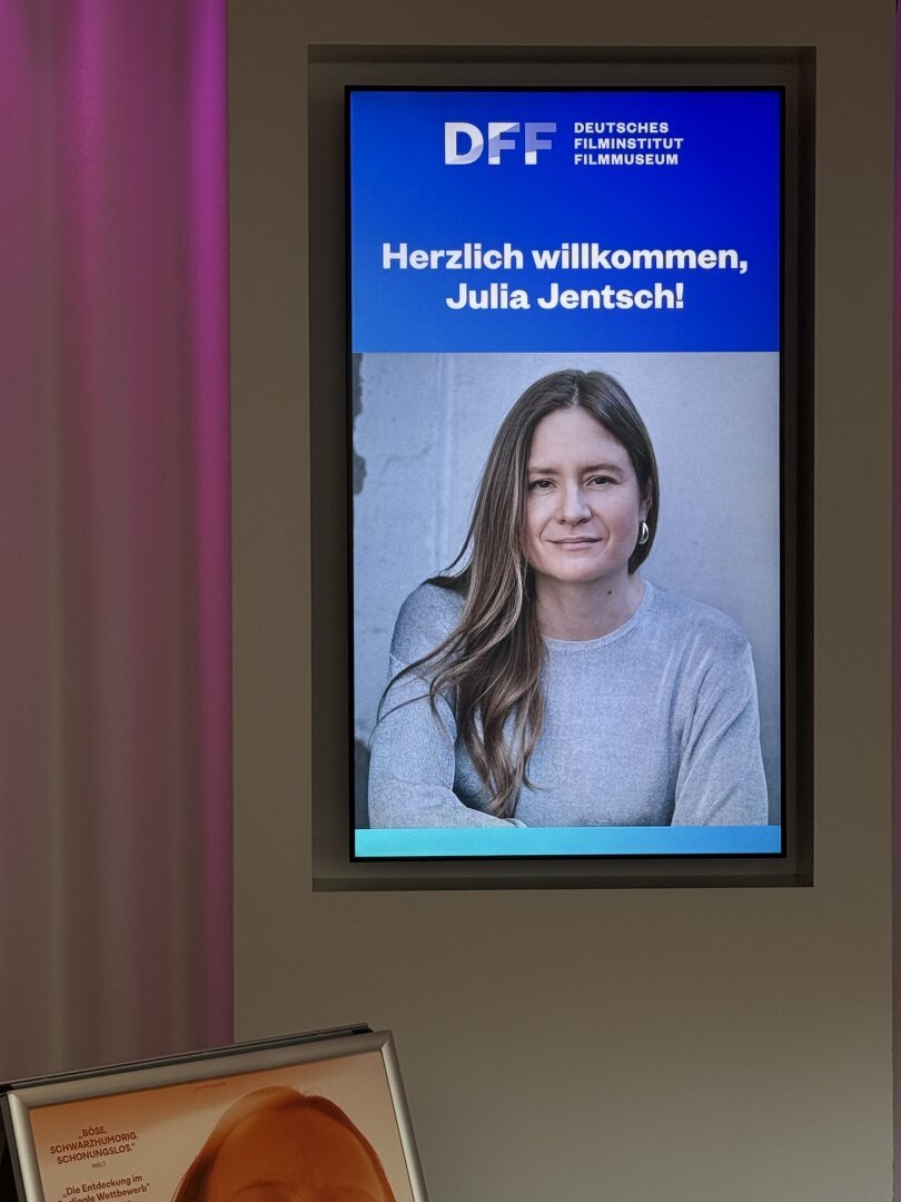 Das Bild zeigt ein digitales Display im Deutschen Filminstitut Filmuseum (DFF). Auf dem Display ist ein großes Foto von Julia Jentsch mit langen, braunen Haaren zu sehen, die ein graues Oberteil trägt. Über dem Foto steht in großen weißen Buchstaben auf blauem Hintergrund: "Herzlich willkommen, Julia Jentsch!" Oben links ist das Logo des DFF zu sehen, gefolgt von der Aufschrift "DEUTSCHES FILMINSTITUT FILMMUSEUM". Im Vordergrund ist ein gerahmtes Bild zu sehen, das einen Ausschnitt eines Films zeigt, mit Text in deutscher Sprache. Die Umgebung ist modern und stilvoll, mit einem leichten Farbverlauf in Violett auf der linken Seite.

Bereitgestellt von @altbot, privat und lokal generiert mit Ovis2-8B
