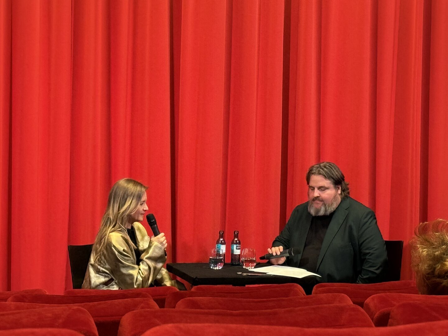 Das Bild zeigt zwei Personen, Julia Jentsch und einen Mitarbeiter des Filmmuseums, die auf einer Bühne sitzen, vor einem roten Vorhang. Die Person links, eine Frau mit langen blonden Haaren, hält ein Mikrofon in der Hand und scheint zu sprechen. Sie trägt eine goldene Jacke. Die Person rechts, ein Mann mit Bart und langen Haaren, sitzt ebenfalls mit einem Mikrofon in der Hand und scheint aufmerksam zuzuhören. Auf dem Tisch zwischen ihnen stehen zwei Flaschen und ein Glas. Im Vordergrund ein paar Sitzreihen des Kinosaals.
Aufgenommen beim fast einstündigen Gespräch vor dem Film.

Bereitgestellt von @altbot, privat und lokal generiert mit Ovis2-8B, etwas überarbeitet.