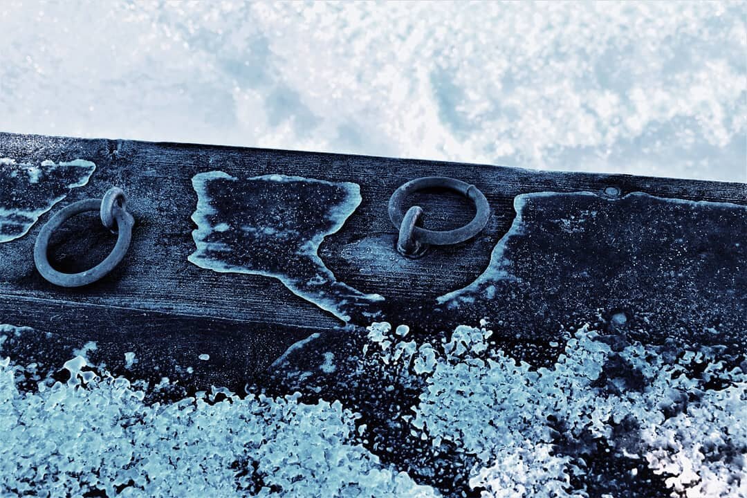 Close up photo of the edge of a wooden dock, two small iron loops are in the wood, theres ice and frost on the wood and below it on the upper part of the photo there's snowy frozen water