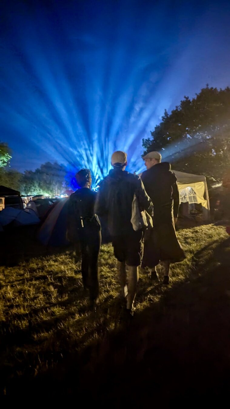 3 Personen gehen in einem Festivalsetting bei Nacht  auf ein Licht zu