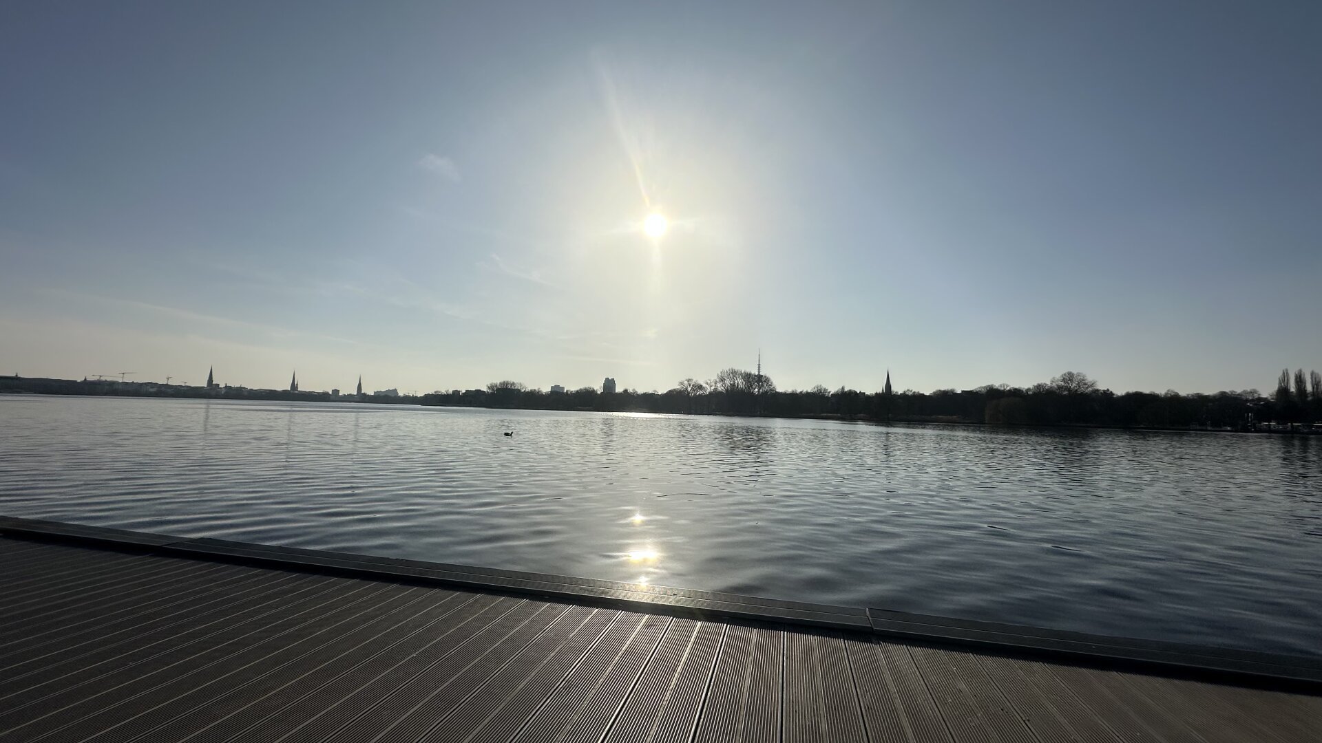 Ein Steg mit Wasser (Alster) in Hamburg. In der Mitte des Bildes die Sonne wie sie auf die Skyline in Hamburg scheint.