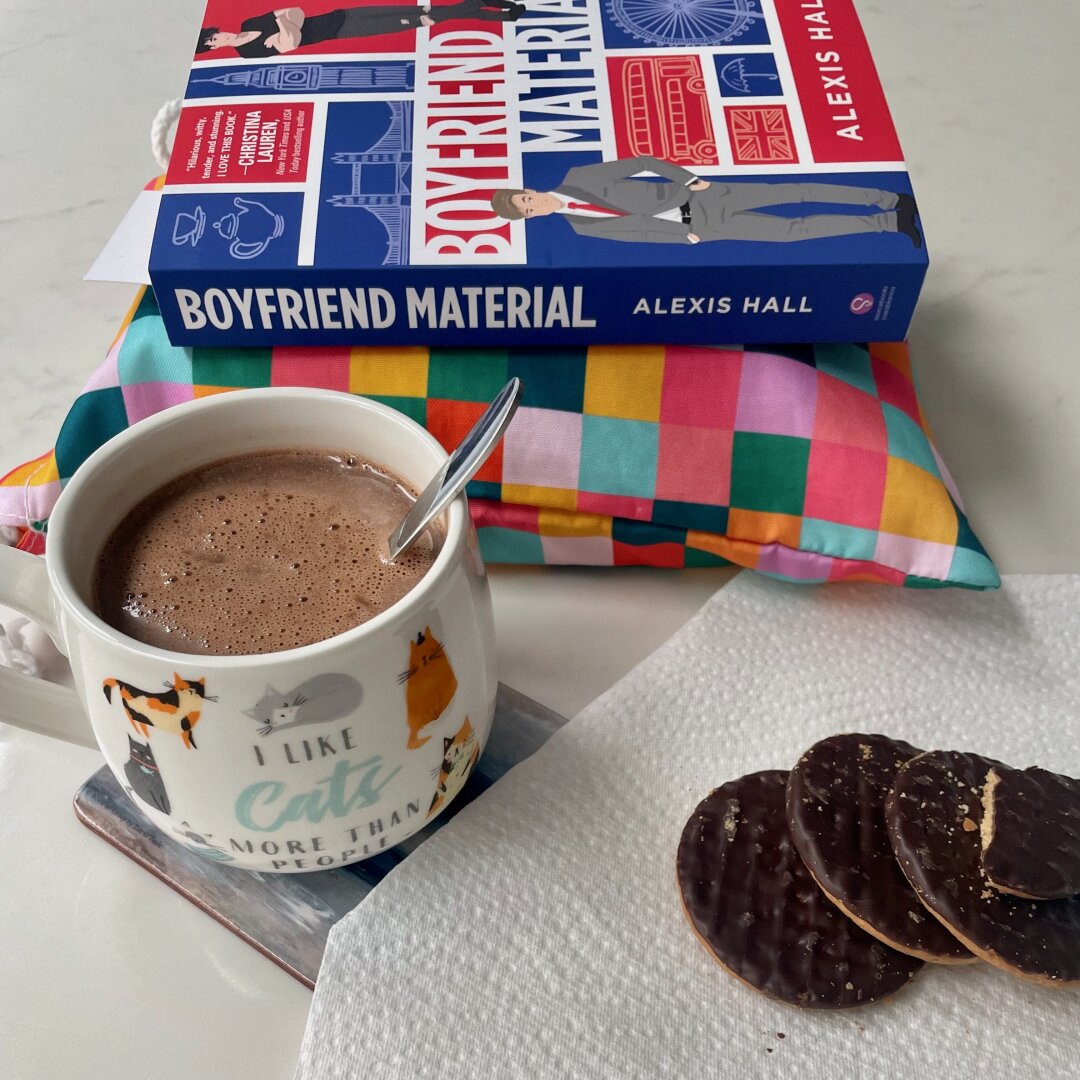 A paperback book is shown alongside a cup of hot chocolate and some dark chocolate digestive biscuits.