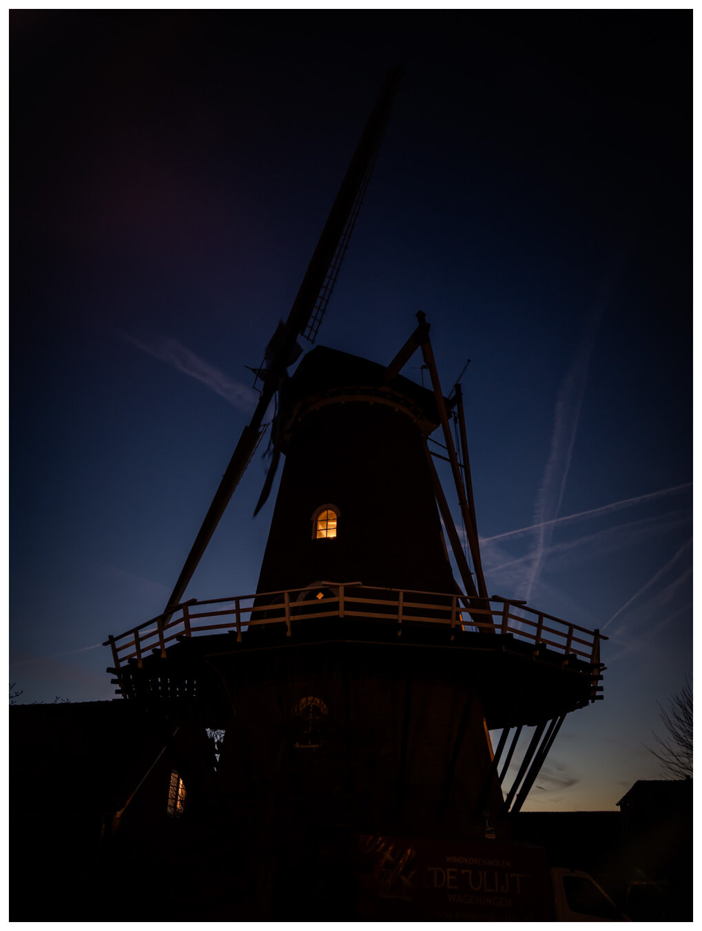 Dark silhouet of a windmill, with one window lighting up. The sky is dark, showing a dark gradient from almost black via clear blue to yellow at the horizon.