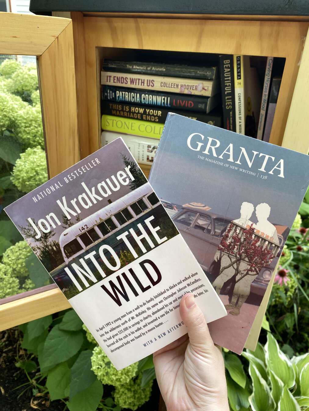 Hand holding up two books in front of a Little Free Library box. The books are Jon Krakauer's Into the Wild and Granta magazine issue 138.