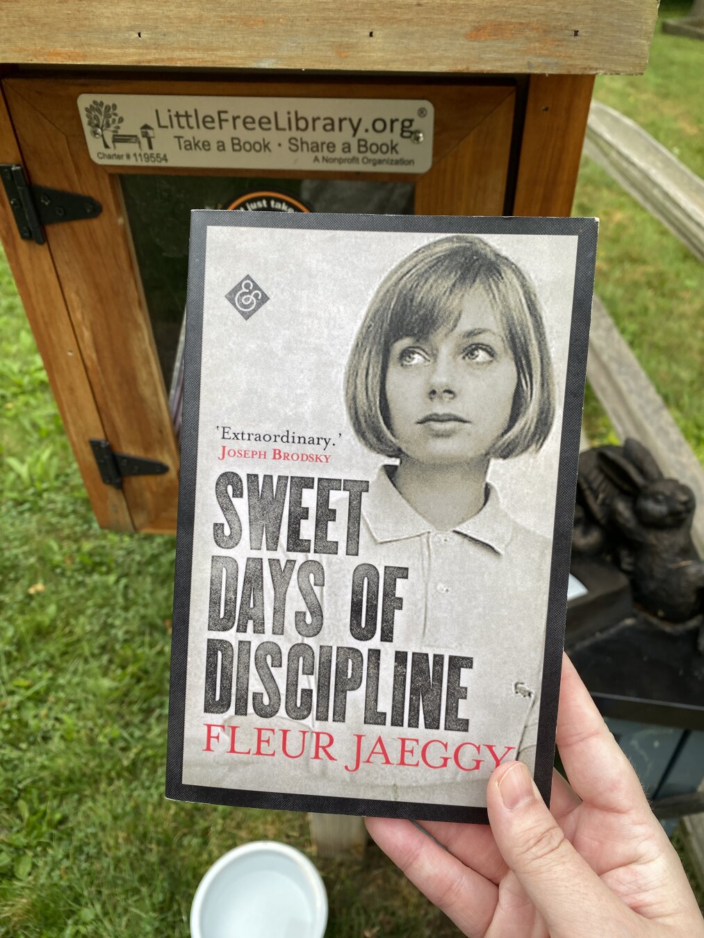 Hand holding up a book in front of a Little Free Library box. The books is Fleur Jaeggy's Sweet Days of Discipline.