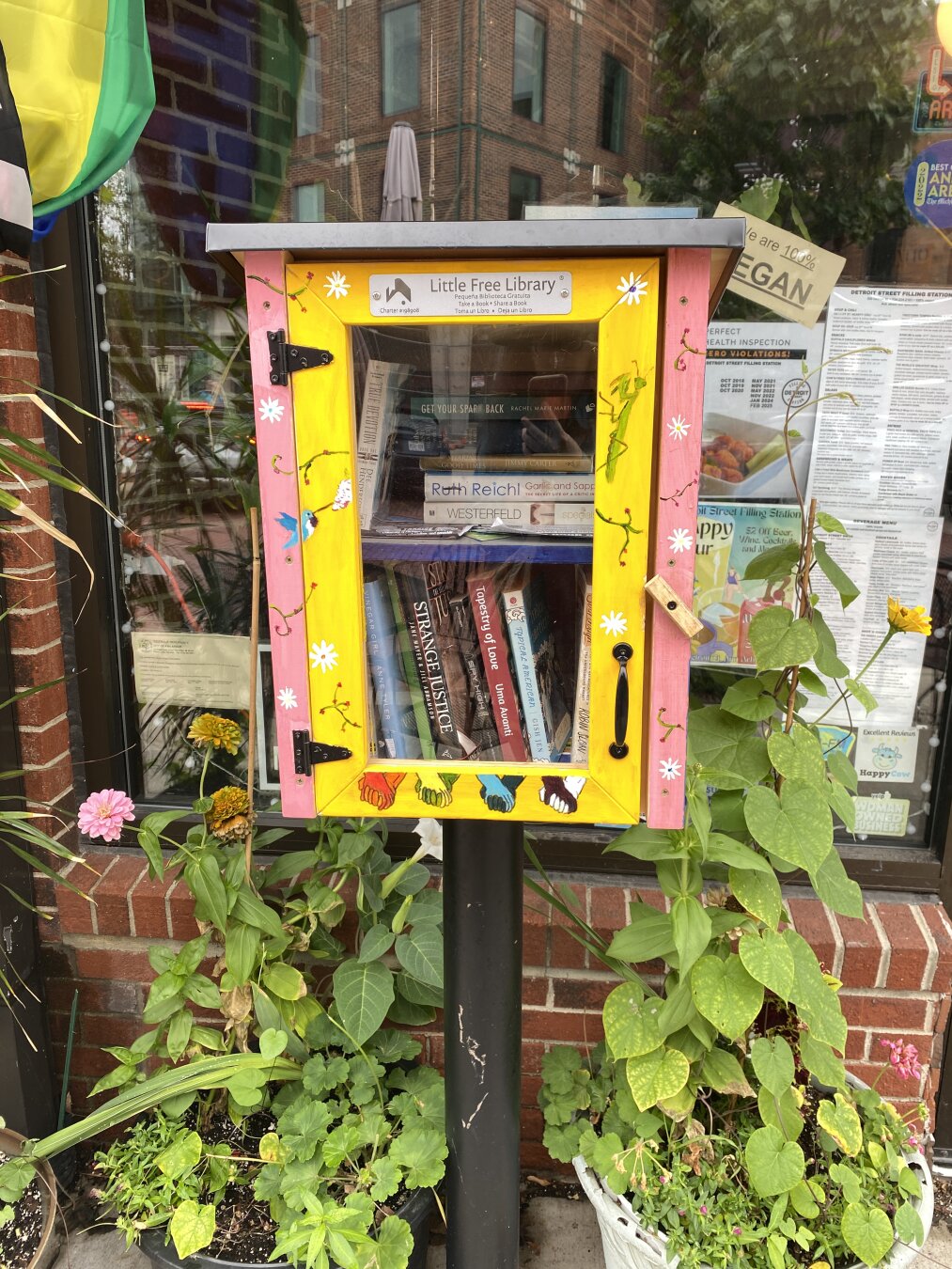 Little Free Library charter #198908 , which has a pink base and a bright yellow door, upon which are drawings showing hands of different colors clasping.