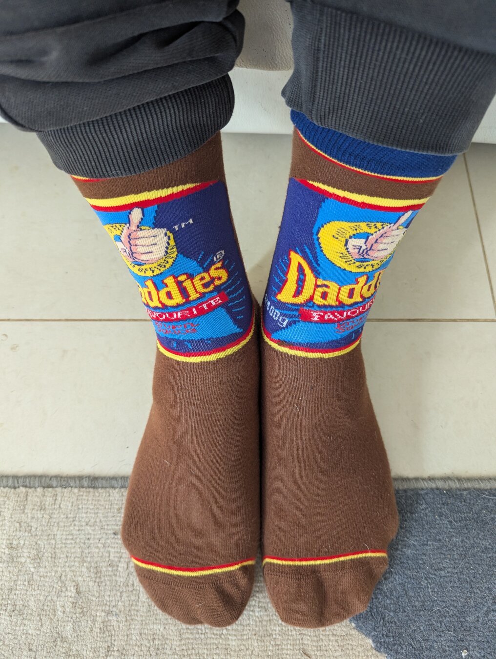 A photo of a person's feet wearing socks. The socks are brown and have a design that is like the label on Daddies Favourite Brown Sauce