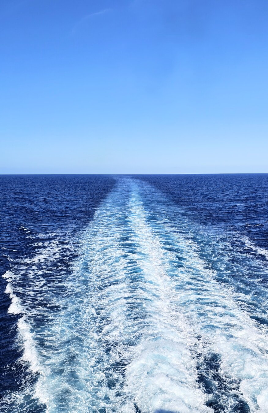 Scia bianca di una nave che si estende verso l'orizzonte su acqua blu scura, cielo sereno azzurro, onde ai lati.