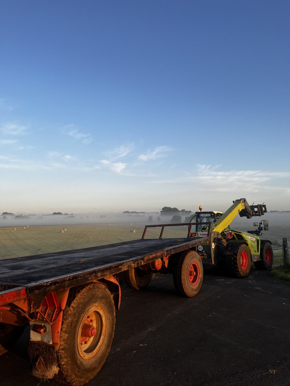 Teleskoplader mit Anhänger von Sonne beschienen, im Hintergrund Fläche mit Heuballen