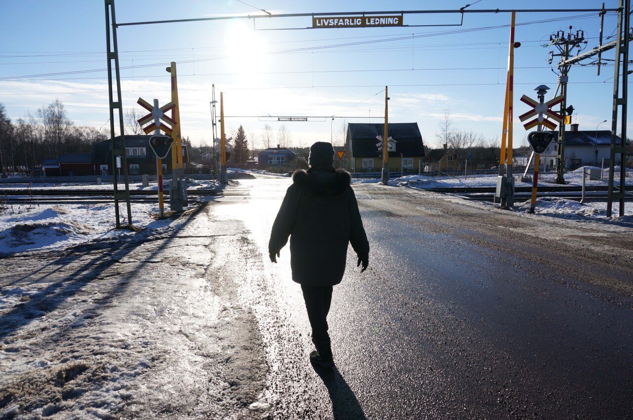 A colour photo of a person walking with their back to camera. They are going towards a train crossing, with the sun in their face.