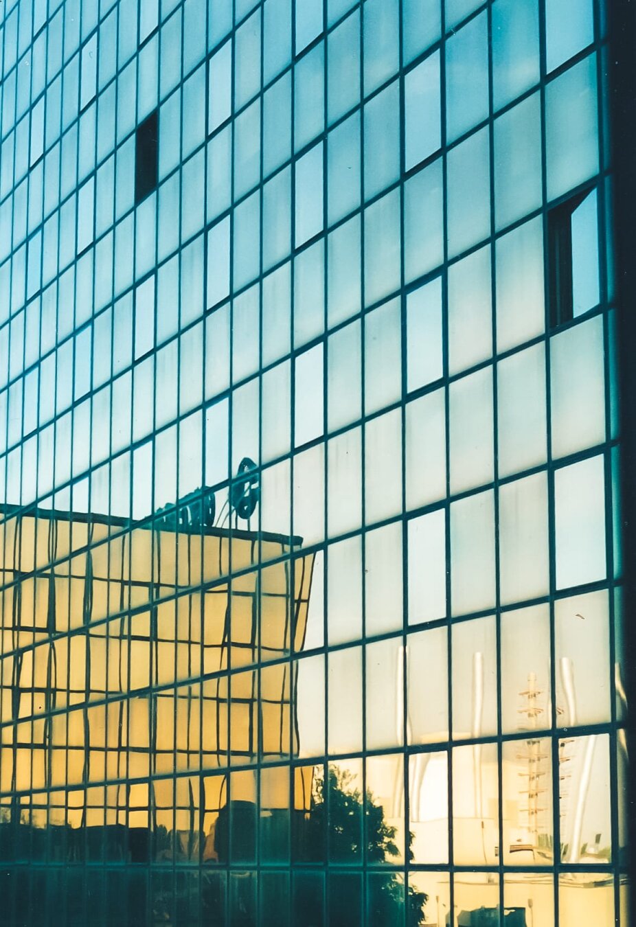The frontage of an office building, so an almost entirely uniform shot of windows (there are a couple open) with the reflection of another building on the left, some trees along the bottom and some industrial chimneys on the right