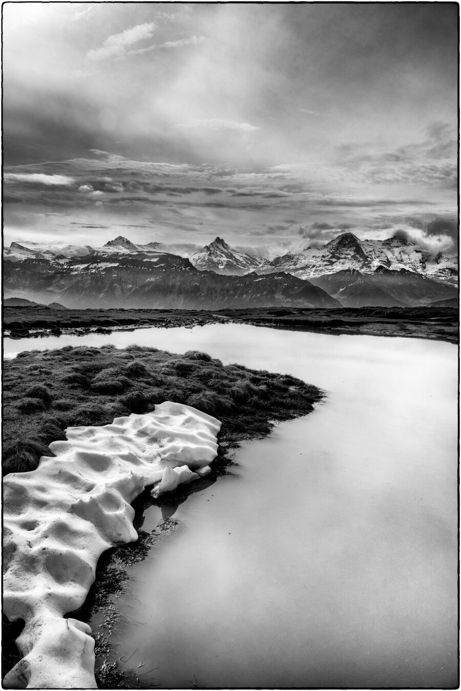 Schwarzweissfotografie im Hochformat eines Teichs mit Schneeresten am Rand. Im Hintergrund sind die Berner Alpen zu sehen. Schwarzweissfotografie im Hochformat eines Teichs mit Schneeresten am Rand. Im Hintergrund sind die Berner Alpen zu sehen.