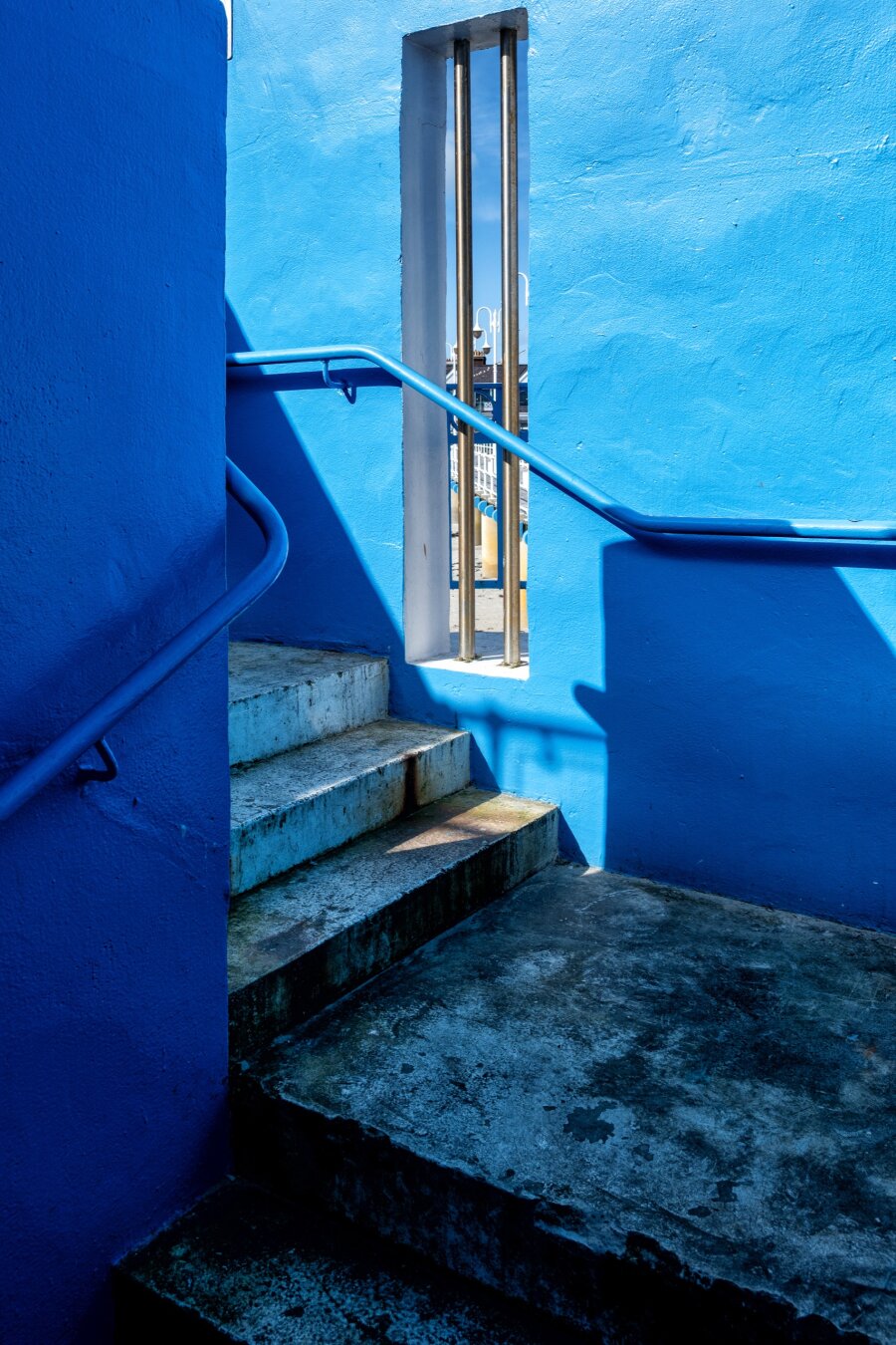 Blauer Treppenaufgang mit metallischem Geländer, der zu einem hellblauen, rechteckigen Ausschnitt in einer Wand führt. Die Treppen sind leicht abgenutzt und zeigen Spuren von Wetter und Zeit. Klare, kräftige Blautöne dominieren die Szene. Foto beschnitten, damit kein Himmel sichtbar ist.