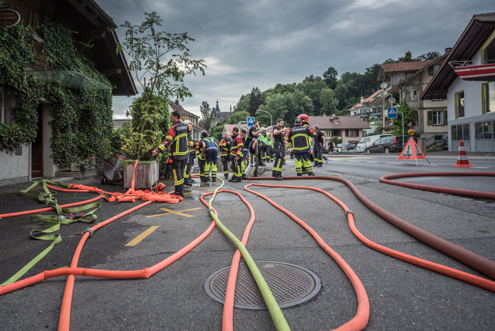 Mehrere Angehörige der Feuerwehr stehen während einer Übung auf einer asphaltierten Strasse und dem angrenzenden Trottoir. Im Vordergrund liegen zahlreiche ausgelegte, leuchtend orangefarbene und gelbe Feuerwehrschläuche geschlängelt auf dem Boden. Links befindet sich ein mit Efeu überwachsenes Gebäude, an dem ein Schild montiert ist. Die Einsatzkräfte in dunkler Schutzkleidung mit gelb-roten Signalstreifen stehen in Gruppen zusammen und besprechen den Einsatz. Rechts im Hintergrund sichern Warndreiecke und ein Leitkegel den Strassenbereich ab, während im Hintergrund Wohnhäuser und ein bewaldeter Hügel unter bedecktem Himmel zu sehen sind.
