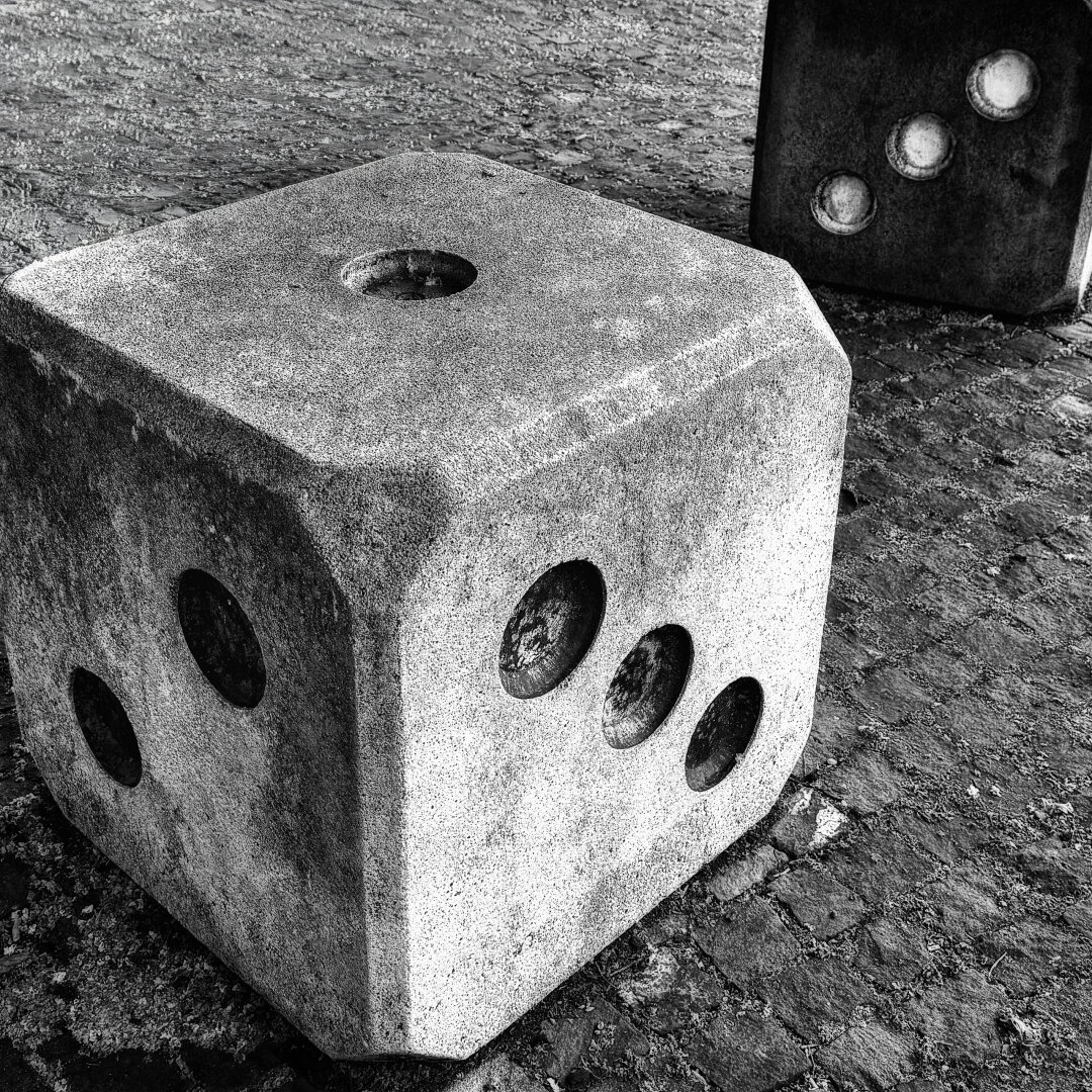 Schwarzweiss-Nahaufnahme von grossen, aus Beton gefertigten Sitzwürfeln im öffentlichen Raum. Im Vordergrund steht ein quadratischer Würfel mit abgerundeten Kanten auf einem Kopfsteinpflaster; sichtbar sind die Oberseite mit einem einzelnen Auge (Eins), die Vorderseite mit drei Augen (Drei) und die linke Seite mit zwei Augen (Zwei). Rechts im Hintergrund ist ein zweiter, im Schatten liegender Steinwürfel zu erkennen, dessen Seite ebenfalls drei Augen zeigt. Die raue Textur des Steins und das Pflaster treten durch den starken Kontrast plastisch hervor.