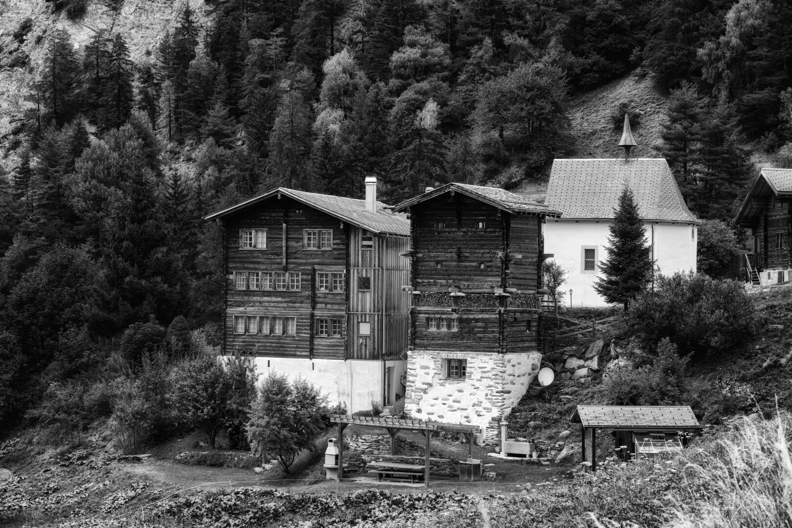 Dunkle, wettergegerbte Holzhäuser mit weissen Sockeln stehen dicht gedrängt an einem steilen, bewaldeten Berghang im Walliser Binntal. Rechts daneben ragt eine kleine, weiss verputzte Kapelle mit spitzem Turm empor, flankiert von einer einzelnen Tanne. Im Vordergrund erstreckt sich ein terrassierter Garten mit einem kleinen Sitzplatz unter einer Pergola, während im Hintergrund dichte Nadelwälder den Hang hinaufwachsen. Die kontrastreiche Schwarzweiss-Ausarbeitung hebt die Texturen des alten Holzes und des Mauerwerks besonders plastisch hervor.