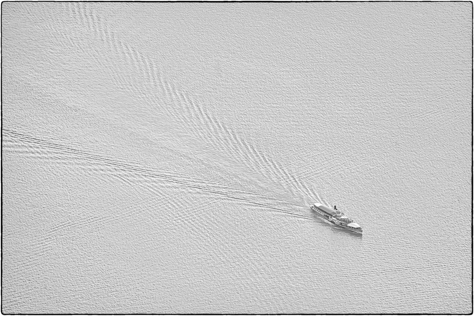 Minimalistische Schwarzweiss-Aufnahme eines Fahrgastschiffs aus stark erhöhter Vogelperspektive. Das kleine Passagierschiff fährt unten rechts im Bild über eine weite, hellgrau schimmernde Wasserfläche. Es zieht eine lange, sich nach oben links diogonal öffnende Kielwelle sowie sanfte Wellenmuster hinter sich her. Die Komposition zeichnet sich durch eine feine Textur der Wasseroberfläche und viel Freiraum um das Schiff aus. Der Übergang wird von einem feinen, dunklen Rahmen eingefasst.