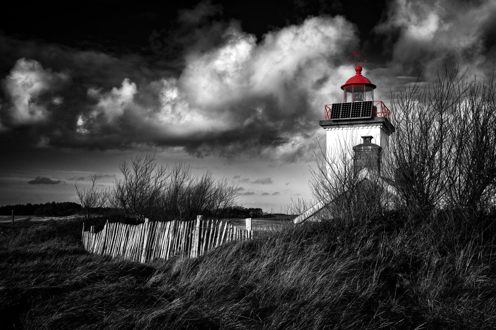Teilkolorierte Schwarzweiss-Aufnahme des Leuchtturms Phare de la pointe d’Agon in der Normandie. Rechts erhebt sich der quadratische, weisse Leuchtturmbau mit einem angrenzenden Gebäudefragment; die Laternenkuppel, das Geländer der Aussichtsplattform sowie die Windfahne heben sich als einziger Farbakzent in leuchtendem Rot ab. An der Galerie der Laterne sind Solarpanels montiert. Im Vordergrund erstreckt sich eine raue Dünenlandschaft mit dunklem Dünengras, kahl verästelten Sträuchern und einem hölzernen Staketenzaun. Ein dramatisch kontrastreicher, dicht mit weissen und dunklen Wolken verhangener Himmel spannt sich über die Szene.