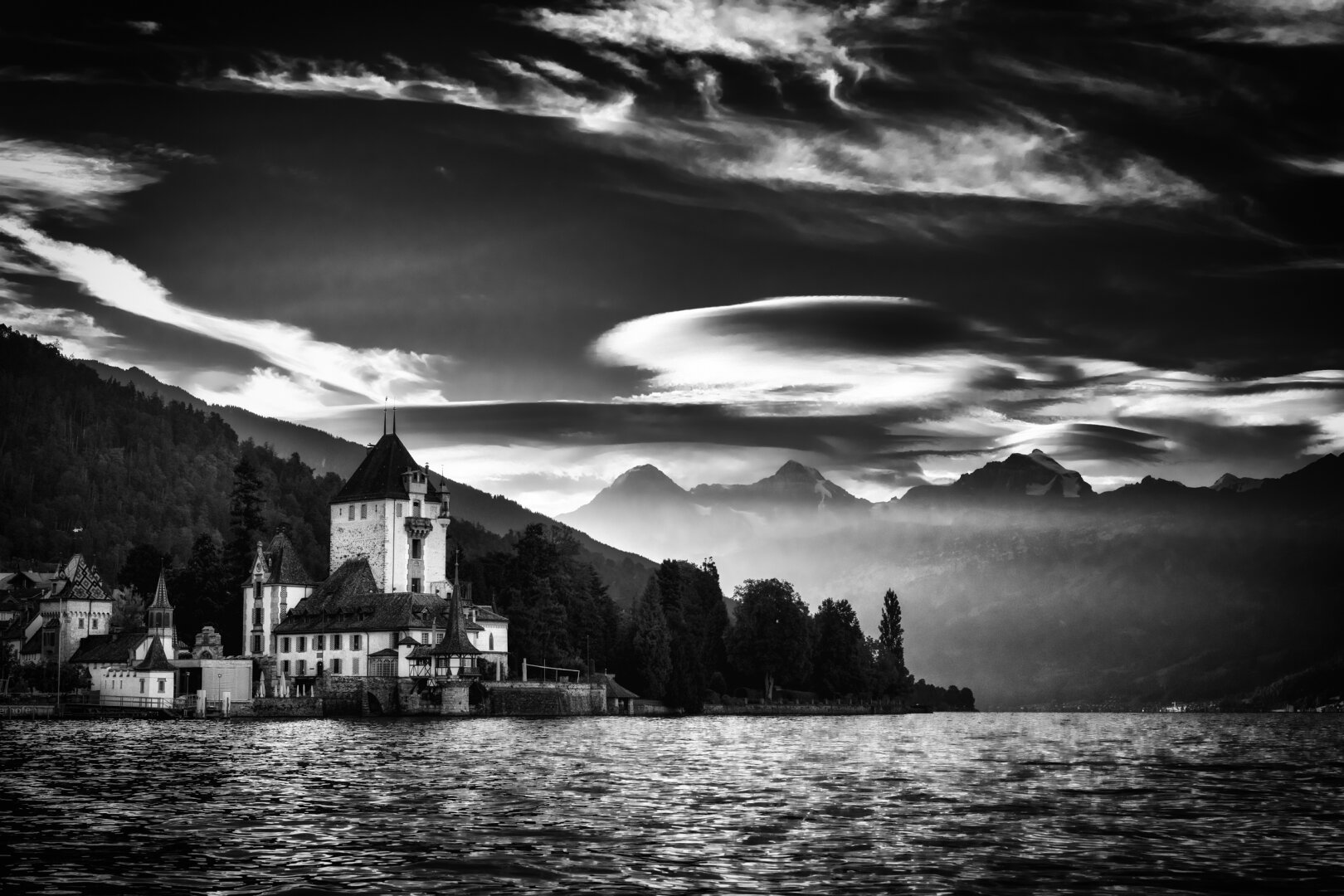 Schloss Oberhofen thront markant am linken Ufer des  Thunersees, während sich im Hintergrund die Gipfel der Berner Alpen aus einer leichten Dunstschicht erheben. Über der alpinen Kulisse spannt sich ein kontrastreicher, dunkler Himmel, der von aussergewöhnlichen, linsenförmigen Lenticularis-Wolken und dynamischen Schleierwolken geprägt ist. Das sanfte Morgenlicht bricht sich auf der leicht gewellten Wasseroberfläche im Vordergrund und hebt die detailreichen Fassaden und Türme der Schlossanlage hervor. Diese monochrome Komposition fängt die mystische und kraftvolle Stimmung eines Schweizer Spätsommermorgens in dramatischen Graustufen ein.