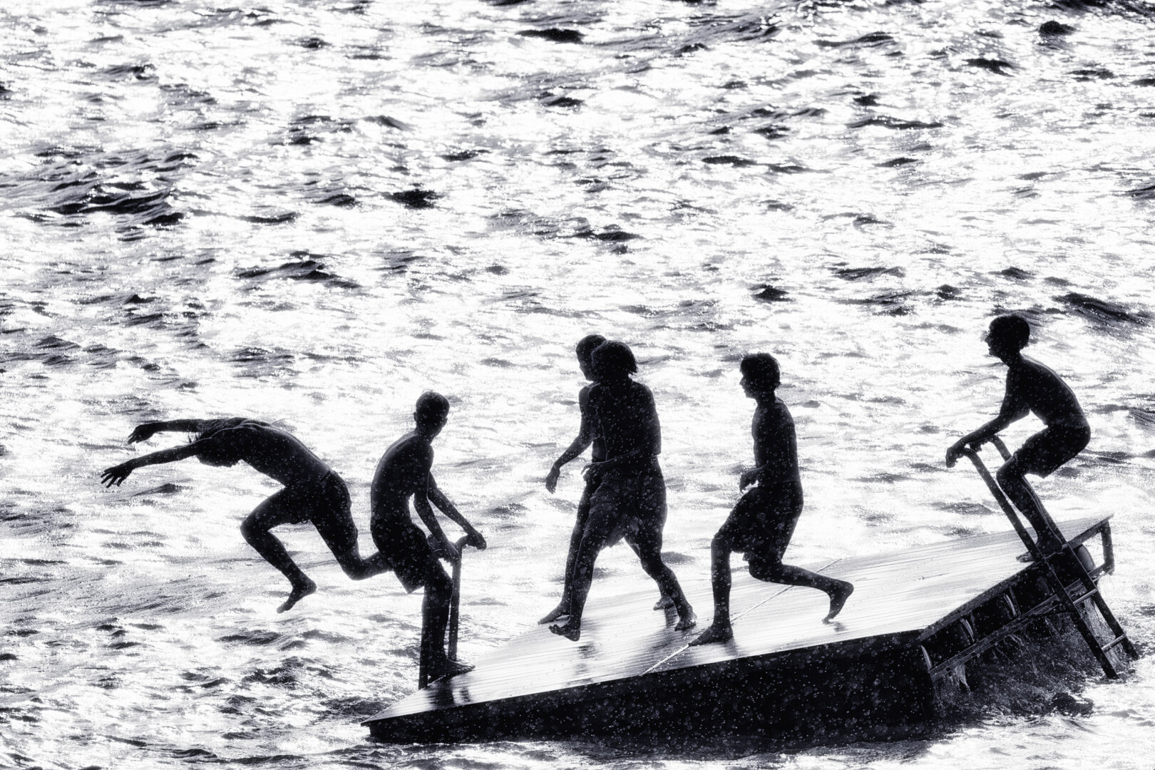Sechs jugendliche Silhouetten bewegen sich dynamisch auf einem schwimmenden Holzfloss inmitten einer stark kontrastierten, glitzernden Wasserfläche. Ein Junge springt mit ausgestreckten Armen schwungvoll ins Wasser, während die anderen auf der Plattform balancieren oder sich an den Griffen festhalten.