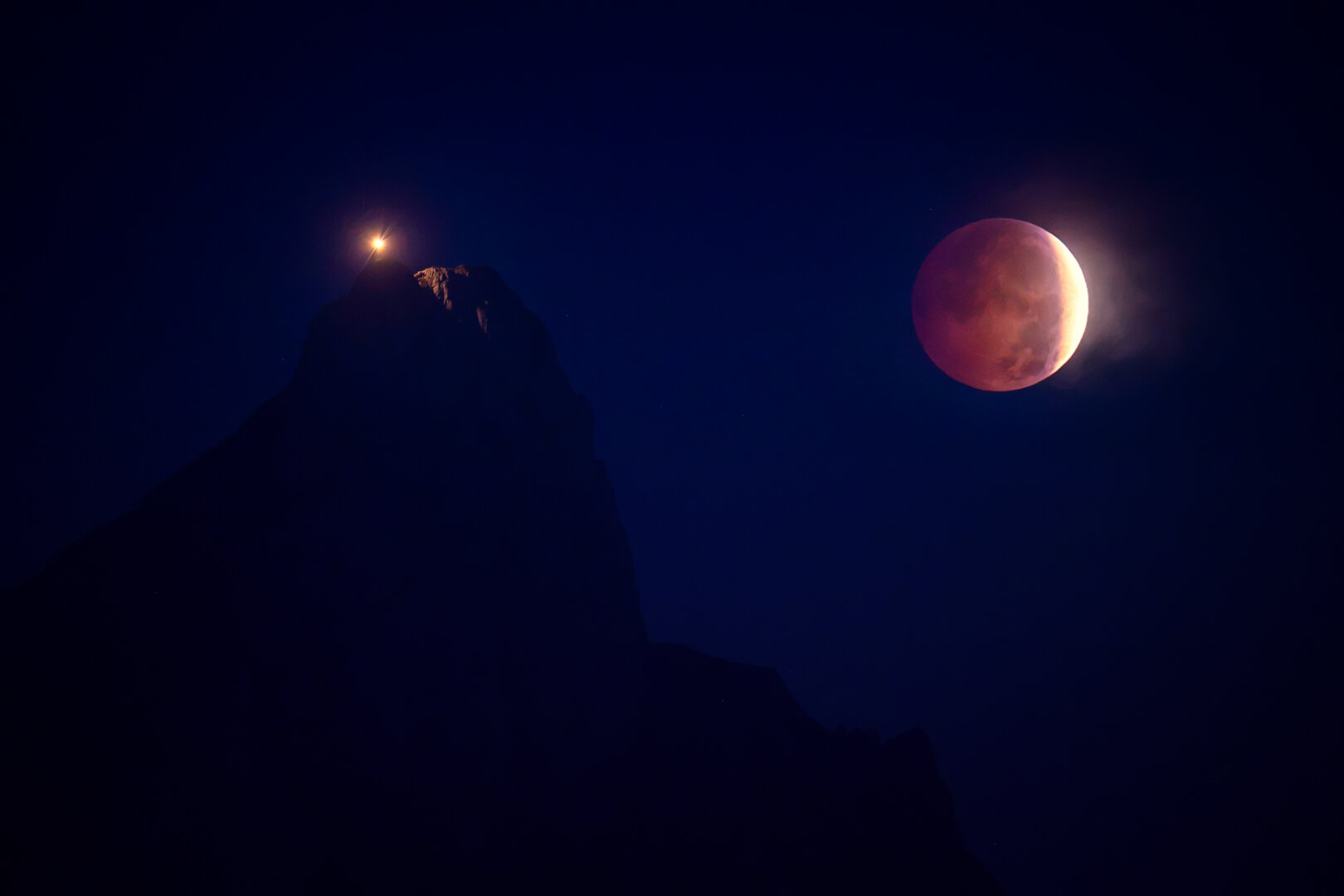 Nächtlicher, tiefblauer Himmel mit einem teilweise verfinsterten, rötlich schimmernden Mond rechts sowie links der Silhouette eines Berggipfels mit einem sternförmig strahlenden Lichtpunkt.