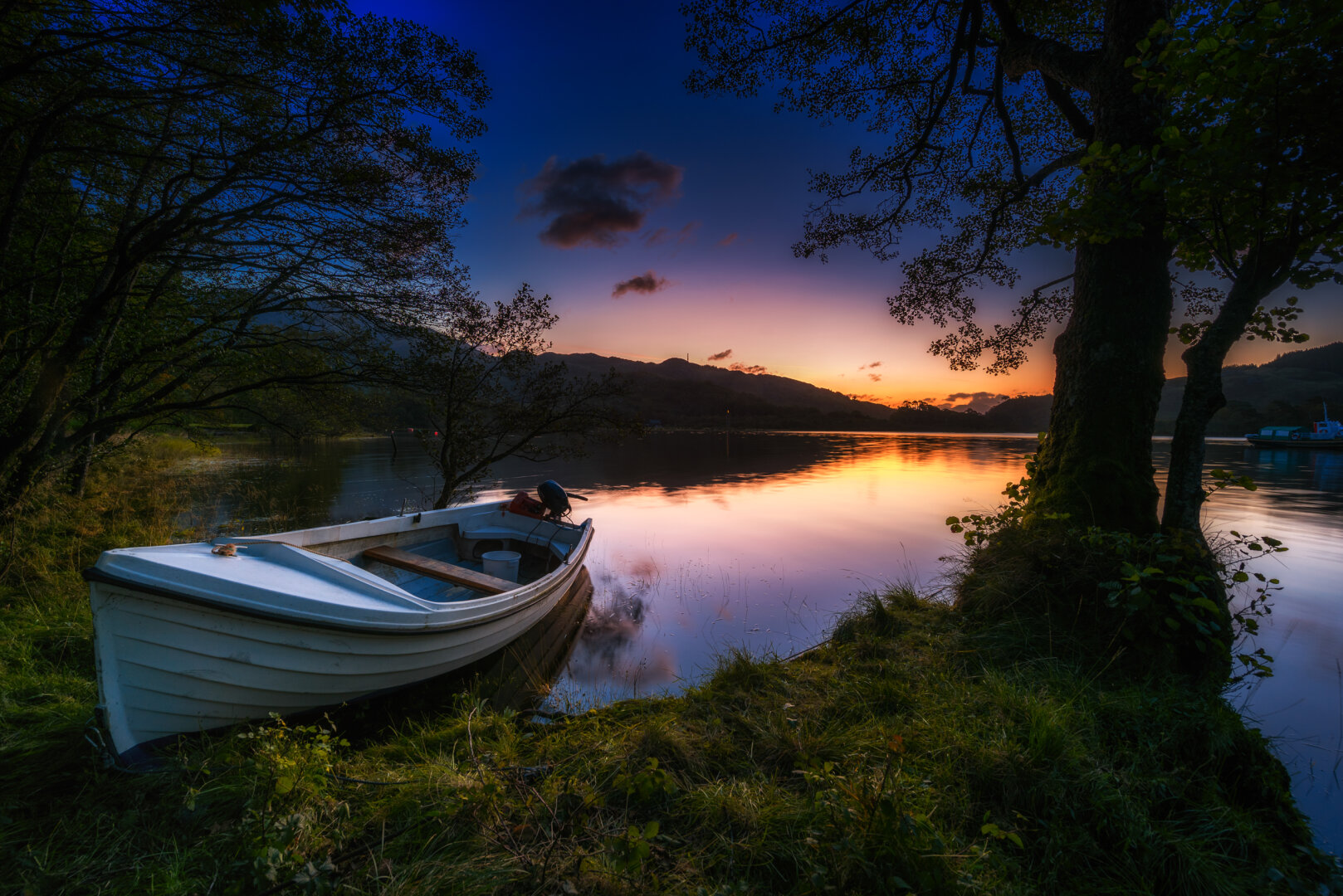 Ein ruhiges, weites Landschaftsfoto aufgenommen am Loch Shiel in den schottischen Highlands in der Morgendämmerung. Ein kleines, weisses Ruderboot liegt am linken Ufer, der Bug ragt leicht aus dem Wasser auf die Uferböschung aus Gras. Der See spiegelt den klaren Himmel wider, der von tiefem Blau im Zenit über ein dunkles Grau in einen leuchtenden, warmen Orange- und Rosaton am Horizont übergeht, wo die Sonne gerade aufzugehen beginnt. Die dunklen Umrisse von Hügeln und Bergen bilden die Horizontlinie. Am rechten Ufer steht ein einzelner, knorriger Baum, der den Rahmen dominiert. Die Szenerie ist friedlich und von weichem, diffusem Morgenlicht erfüllt.