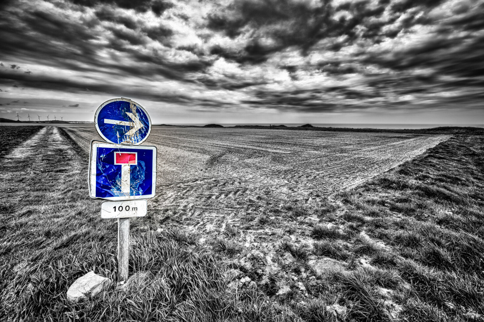 In einer kargen, monochrom gehaltenen Agrarlandschaft steht am linken Bildrand ein hölzerner Pfosten mit drei beschädigten Verkehrsschildern. Das oberste runde Schild zeigt einen blauen Hintergrund mit einem weissen, nach rechts gerichteten Pfeil. In der Mitte befindet sich ein rechteckiges Schild, das eine Sackgasse anzeigt: auf blauem Grund ist ein weisses T mit einem roten Querbalken abgebildet, darunter  ein kleines Zusatzschild mit der Aufschrift „100 m“. Die Schilder sind stark zerkratzt und verbeult. Der Hintergrund wird von einem flachen, frisch gepflügten Feld und einer fernen Horizontlinie eingenommen. Darüber spannt sich ein dramatischer, tief hängender Wolkenhimmel. Die gesamte Szene ist bis auf das Blau und den roten Balken der Schilder  in kontrastreichen Grau- und Schwarzstufern gehalten.
Diese Fotografie ist als FineArt Druck in einer limitierten Auflage erhältlich http://art.hess.photography/161-dead-end.html