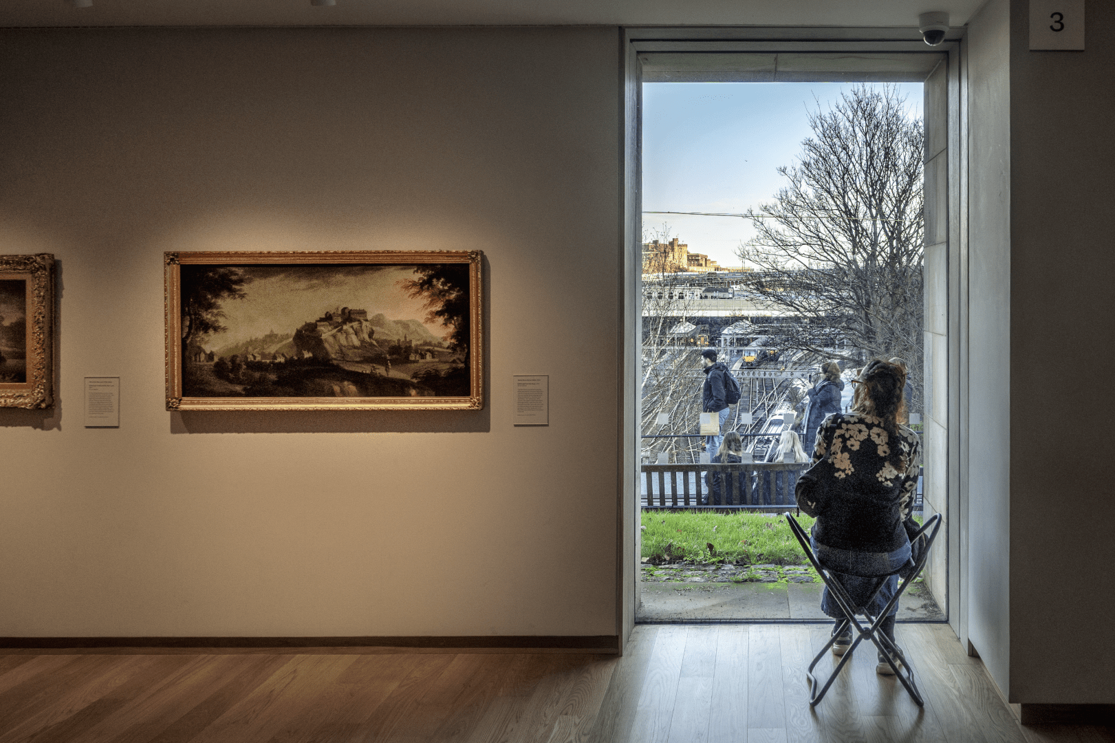 Ein lichtdurchfluteter, moderner Ausstellungsraum mit hellem Holzboden und weissen Wänden. An der linken Wand hängen klassische Gemälde in goldfarbenen Rahmen. Rechts öffnet sich ein hohes, bodentiefes Fenster mit Blick ins Freie. Im Vordergrund sitzt eine Person von hinten gesehen auf einem dunklen Klappstuhl vor der Scheibe. Draussen ist ein grüner Grasstreifen zu sehen, dahinter ein Geländer, an dem mehrere Menschen stehen. Der Blick schweift über Gleisanlagen mit Zügen und einen unbewillten Baum ohne Laub hin zu historischen Gebäude- und Festungsstrukturen unter leicht bewölktem Himmel.