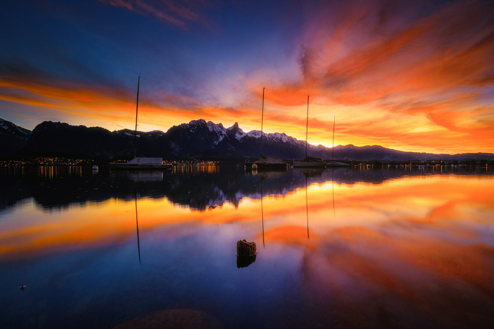 Stimmungsvolle Fotografie eines Sees während eines intensiven Abendrots. Der Himmel leuchtet in kräftigen Orange-, Rot- und Blautönen mit leichten Schleierwolken. Im Hintergrund zeichnet sich die dunkle Silhouettierung einer Bergkette ab, vor der mehrere Segelboote auf dem ruhigen Wasser anlegen. Die farbenprächtige Himmelsstimmung und die Masten der Boote spiegeln sich fast spiegelglatt auf der Wasseroberfläche wider, während im Vordergrund ein kleiner Holzpflock aus dem Wasser ragt.