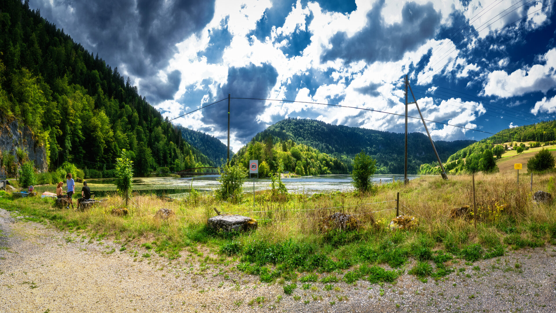 Panoramablick über einen von Wald und Hügeln gesäumten Fluss mit einer Brücke im Hintergrund, im Vordergrund eine Wiese mit Kies-/Schotterweg, Strommasten und drei sitzenden Personen unter dramatisch bewölktem Himmel.