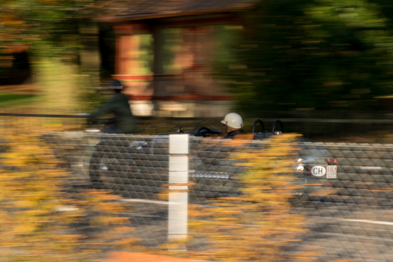 Bewegungsunscharfe Aufnahme eines Oldtimers mit Fahrer im weissen Helm, im Hintergrund ein Radfahrer und ein Gebäude, umgeben von herbstlich gefärbtem Laub.