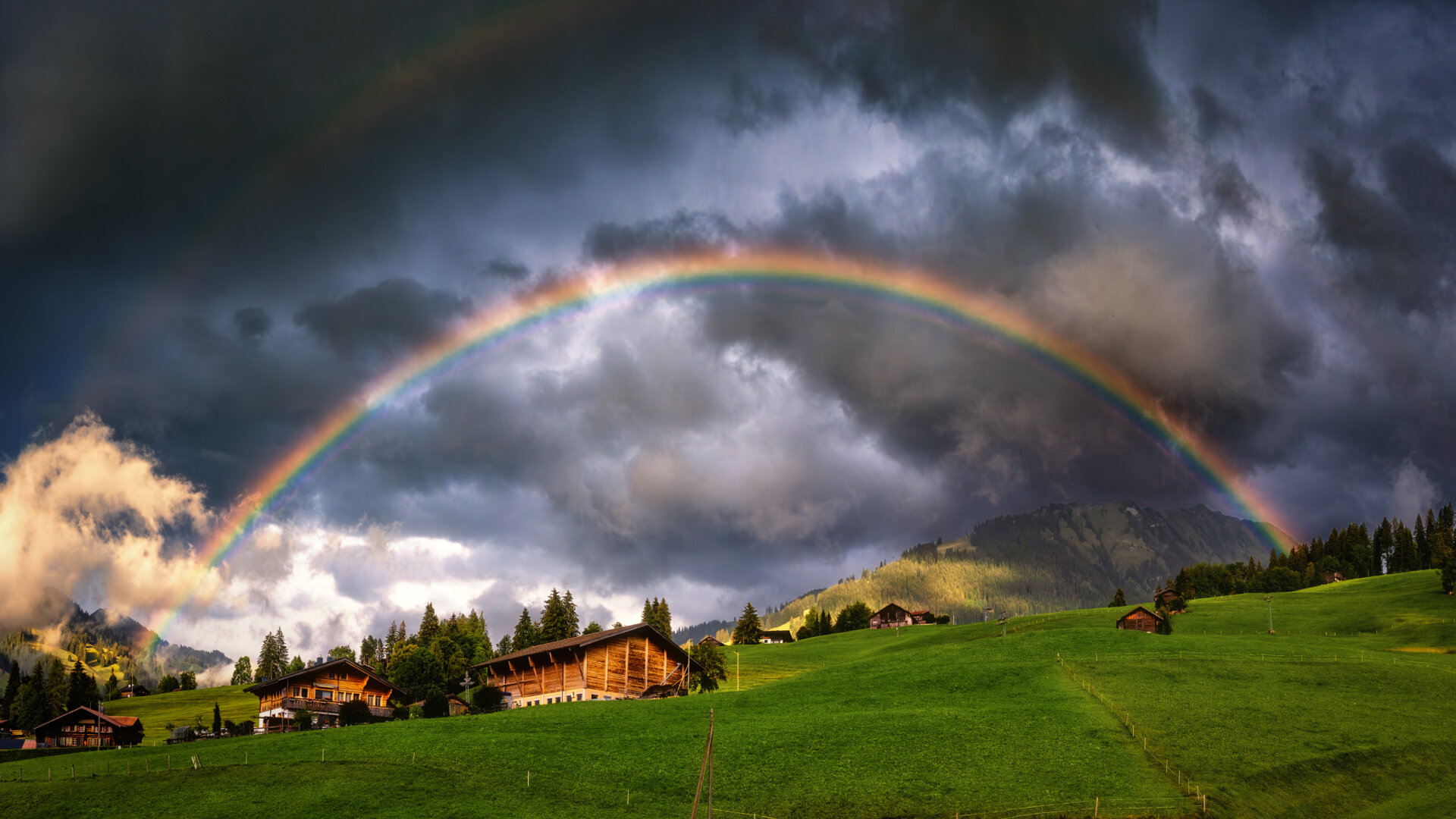 Ein leuchtender, vollendeter Regenbogen spannt sich in einem weiten Bogen über eine hügelige, sattgrüne Alplandschaft mit vereinzelten Holzhütten und Chalets. Im Hintergrund ragen bewaldete Berghänge unter einer dramatischen, dunkelgrauen Gewitterwolkenwand empor, die stellenweise von goldenem Sonnenlicht durchbrochen wird. Das warme Licht der tiefstehenden Spätsommersonne beleuchtet die grünen Wiesen im Vordergrund und lässt die nassen Holzfassaden der Häuser sanft schimmern.