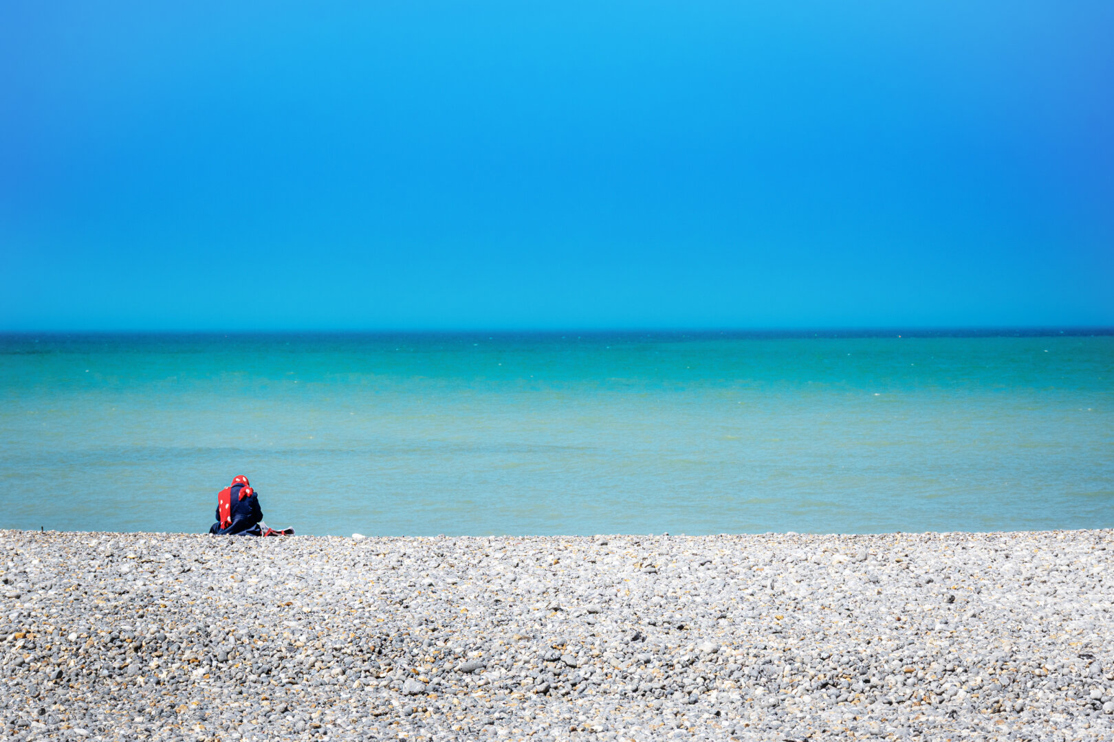 Eine einzelne Person in einer dunkelblauen Jacke und einem auffälligen, rot-weiss gepunkteten Kopftuch sitzt abgewandt auf einem breiten, hellgrauen Kieselstrand. Vor ihr erstreckt sich das ruhig daliegende Meer, das in sanften Türkis- und Blautönen schimmert und am Horizont nahtlos in einen wolkenlosen, tiefblauen Himmel übergeht. Die minimalistische Dreiteilung des Raumes aus grauem Stein, türkisem Wasser und blauem Himmel erzeugt eine Atmosphäre von tiefer Ruhe und Einsamkeit. Das helle Mittagslicht des Frühlingstages lässt die Farben intensiv leuchten und wirft weiche Schatten auf die runden Kieselsteine.